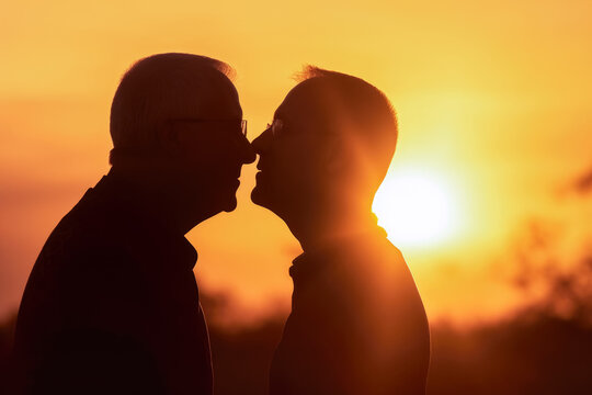 Two men older gay couple kissing at sunset, closeup detail to faces, orange sky behind them. Happy candid romantic moment - LBGT concept. Generative AI