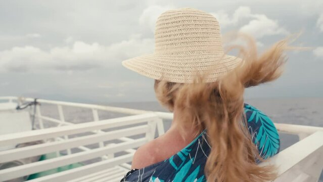 Anonymous blonde woman with long hair and straw hat sitting on upper deck of ferry ship motorboat enjoying freedom and wind in open sea. Female elderly person on ocean cruise voyage on summer vacation