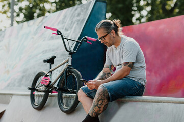 A serious urban tattooed middle-aged man is sitting in a skate park next to his bmx bike and scrolling on his phone.