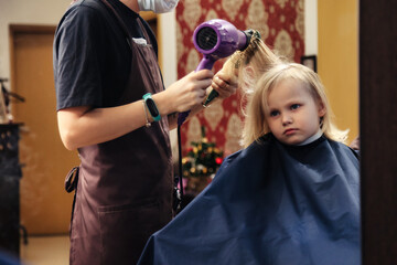 Hair salon, hairdresser dries hair with hair dryer for kid in barber shop at mirror. Barber woman...