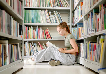 Girl teenager sitting in library on floor and reading book indoors. Beautiful student preparing for exam with education mterials near bookshelfs