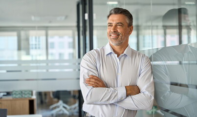 Happy mid aged older professional business man, mature latin executive manager, 50 years old investor standing in office arms crossed leaning at glass wall looking away thinking of success concept.