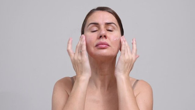 Middle Aged Woman Grey Background. Female 40 Year Lady Applying Moisturizing Cream Face. Facial Massage Enjoying Her beauty, High Self Esteem, Wellness. Natural Cosmetic Skincare Products.