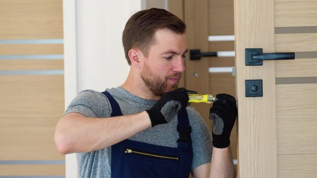 Installation of a lock on the front wooden interior door. Portrait of young locksmith workman in uniform installing knob. Professional repair service. Maintenance Concept