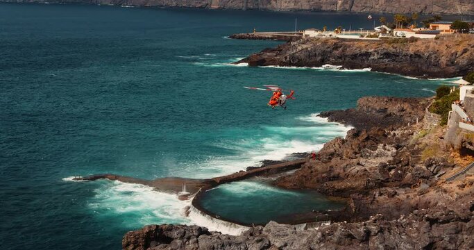 Red rescue helicopter flies over a stormy ocean and lifts a person on board near rocky coastline.