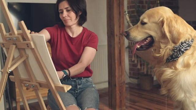 Pretty girl artist makes sketch of golden retriever dog using pencil and canvas at home. Beautiful young woman painting portrait of cute doggy pet