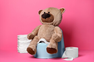 Teddy bear on baby potty, toilet paper and stack of diapers on pink background