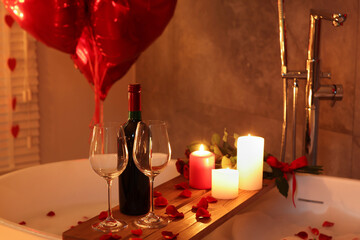Wooden tray with wine, burning candles and rose petals on tub in bathroom. Valentine's day celebration