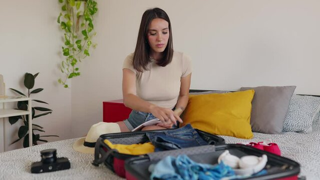 Young beautiful woman sitting on bed packing her suitcase for travel vacation. Smiling female traveler writing planner summer trip on notebook. Summer vacation lifestyle concept.