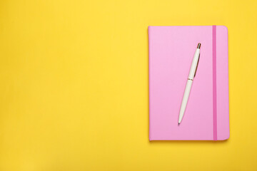 Closed pink notebook and pen on yellow background, top view. Space for text