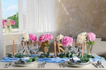 Beautiful table setting. Plates with greeting cards, napkins and branches near glasses, peonies, burning candles and cutlery on table in room