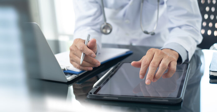 Medicine doctor working on digital tablet and laptop computer searching the information with medical document on table, close up. Doctor writing prescription and filling health insurance document
