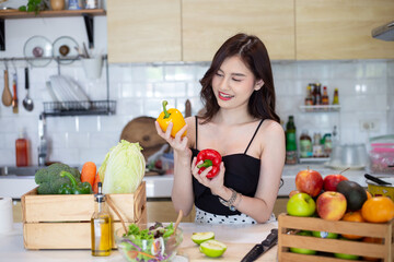 beautiful asian woman smiling preparing, holding a sweet pepper and cut green apple in white kitchen with plenty of fresh vegetables and fruits on the table. At Home