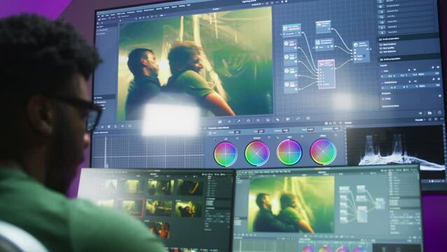 Back view of African American male colorist works at home office, makes color grading. Action movie footage and program interface on computer and big digital screen. Concept of film post production.
