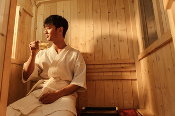 Relaxation Asian man in bathrobe drinking hot tea while relaxing in the sauna. Young male healthcare and spa treatment concept..