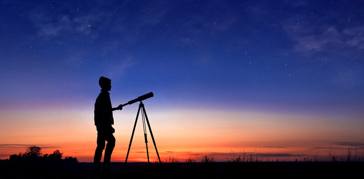 The child looks at the stars through a telescope. A teenager boy looks at the night sky through a spyglass against the backdrop of a sunset while in the field.