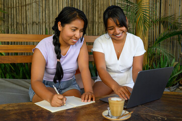 Cheerful latin young women taking notes while working on laptop in cafe