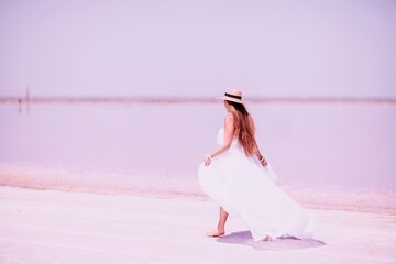 Naklejka premium Woman in pink salt lake. She in a white dress and hat enjoys the scenic view of a pink salt lake as she walks along the white, salty shore, creating a lasting memory.