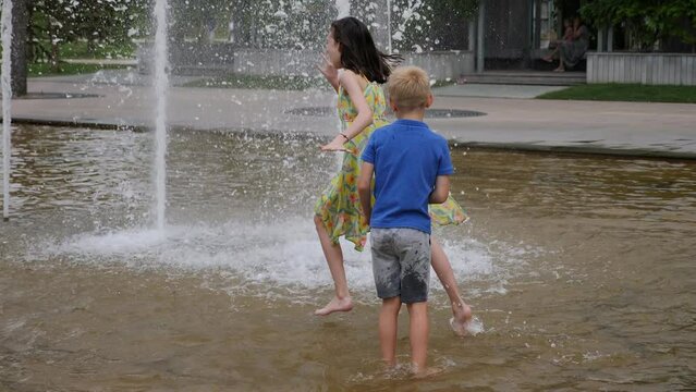Two cheerful children play in the fountain in the city park in the summer. Children in wet clothes play on a hot day in a fountain in a modern city park.