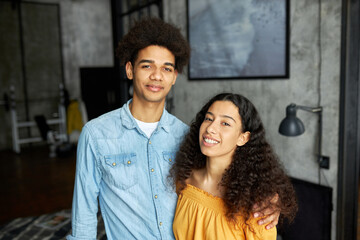 Young married african american couple posing against loft style interior apartment, guy in denim shirt hugging his charming adorable with curly hair, looking at camera. Happy relationships