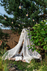 A white net canopy with a chandelier and cushions hangs from a pine tree in the garden