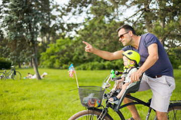 Look over there. Active family day in nature. Father and son ride bike through city park on unny summer day. A cute boy is sitting in front bicycle chair while father rides bicycle. Father son bonding