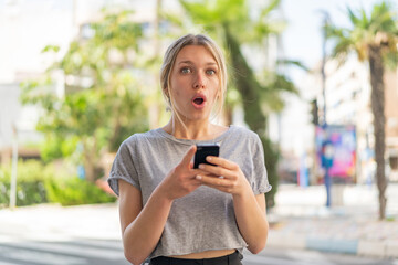 Young blonde woman at outdoors looking at the camera while using the mobile with surprised expression