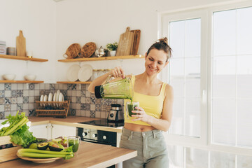 Happy woman on detox diet, pouring green cocktail from mixer into glass in kitchen