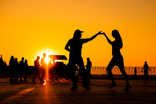 Man and woman couple silhouettes dancing against warm sunset orange sky on quay at evening - sun lens flare. Group dance, romantic, love, summer outdoor activity and vacation concept