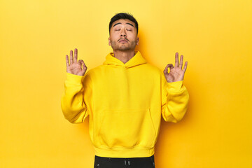 Bold Asian man in yellow hoodie on yellow studio relaxes after hard working day, she is performing...