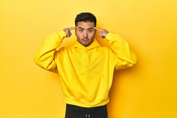 Bold Asian man in yellow hoodie on yellow studio focused on a task, keeping forefingers pointing head.