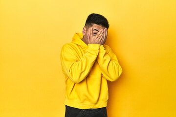 Obraz premium Bold Asian man in yellow hoodie on yellow studio blink through fingers frightened and nervous.