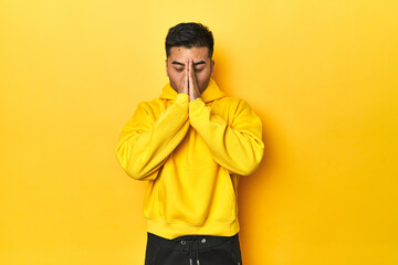 Bold Asian man in yellow hoodie on yellow studio holding hands in pray near mouth, feels confident.