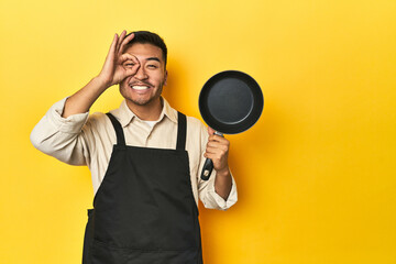 Asian chef holding a pan, yellow studio backdrop excited keeping ok gesture on eye.
