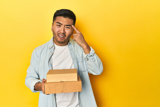 Asian Man Holding Delivery Food Boxes, Yellow Studio Backdrop Showing A Disappointment Gesture With Forefinger.