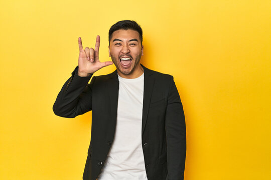 Asian man in black business jacket on yellow studio showing a horns gesture as a revolution concept.