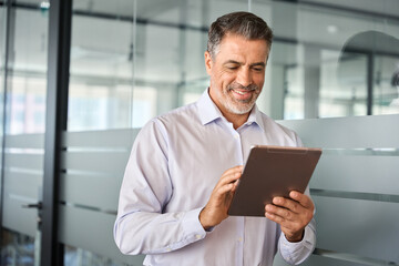 Young smiling busy mature male manager using tablet standing in office. Older mid aged professional...