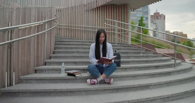 Education in university college concept - young lady reading notebook while sitting outdoors on steps