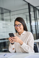Smiling young Asian business woman using smartphone sitting in office. Happy professional Japanese female manager worker wearing glasses holding cell working on mobile phone, vertical.