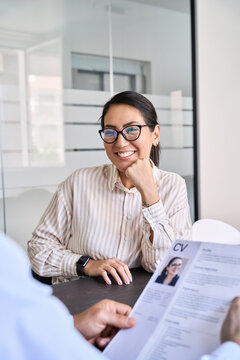 Smiling professional Asian business woman candidate and hr manager employer holding cv resume during job interview sitting in office at meeting. Human resources concept. Over shoulder vertical shot.