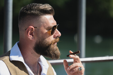 Set portrait. Man smoking a cigar by the river with long beard, shirt and curled moustache.
Dark...
