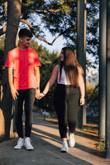Joven y guapa Pareja disfrutando de un día soleado mientras pasean cogidos de la mano por un camino, en un parque con arboles.