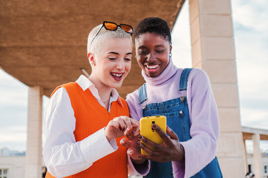 Two happy women friends smiling and watching funny photos on a social media cellphone app. A pair of girls having fun buying together on a online shop with a smart phone. Ladies using a mobile phone