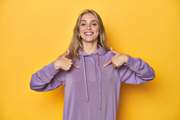 Young blonde Caucasian woman in a violet sweatshirt on a yellow background, surprised pointing with...