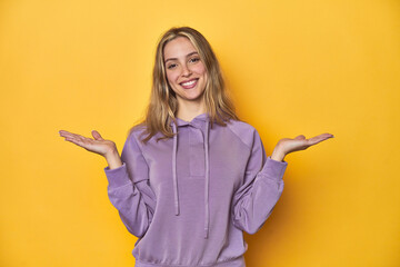 Young blonde Caucasian woman in a violet sweatshirt on a yellow background, makes scale with arms, feels happy and confident.