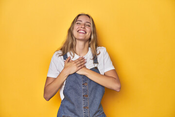 Young blonde Caucasian woman in denim overalls posing on a yellow background, laughing keeping hands on heart, concept of happiness.