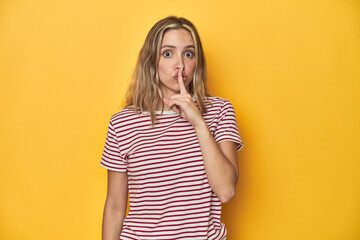 Young blonde Caucasian woman in a red striped t-shirt on a yellow background, keeping a secret or asking for silence.