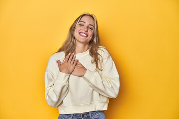 Fototapeta premium Young blonde Caucasian woman in a white sweatshirt on a yellow studio background, has friendly expression, pressing palm to chest. Love concept.