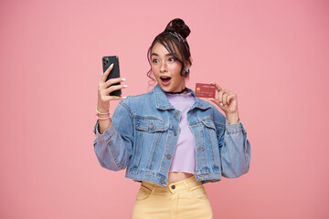 surprised Asian young girl using smartphone and showing credit card isolated on pink background....