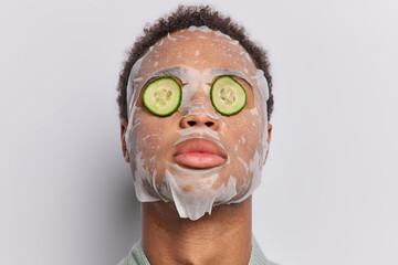 Fototapeta premium Photo of dark haired man applies sheet facial mask covers eyes with cucumber slices undergoes daily procedures for rejuvenation isolated over white background. People wellness and skin care concept